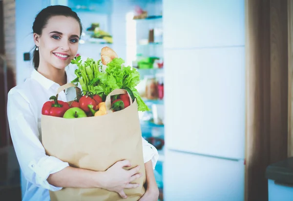 Jeune femme tenant sac d'épicerie avec des légumes. Debout dans la cuisine. Femme dans la cuisine regardant la caméra — Photo