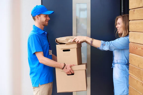 Lachende bezorger in blauw uniform levert pakketdoos aan ontvanger - koerier service concept. Glimlachende bezorger in blauw uniform — Stockfoto