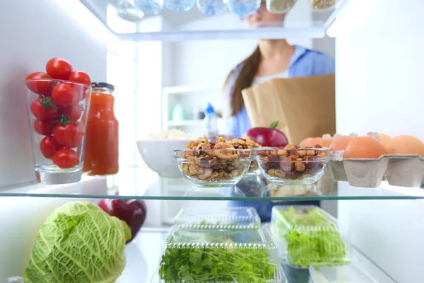 Портрет женщины, стоящей рядом с открытым холодильником, полным здоровой пищи, овощей и фруктов — стоковое фото