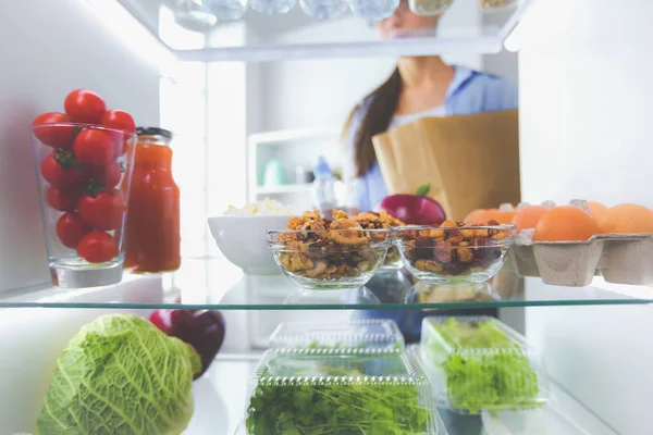 รูปภาพของผู้หญิงยืนอยู่ใกล้ตู้เย็นเปิดเต็มไปด้วยอาหารเพื่อสุขภาพผักและผลไม้ — ภาพถ่ายสต็อก