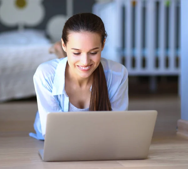 Ung kvinna som sitter på golvet nära barnens babysäng med laptop. Ung mamma — Stockfoto