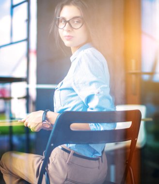 Ofis masasında dizüstü bilgisayarıyla oturan genç bir kadın. Pencereden bakıyor. Genç kadın.