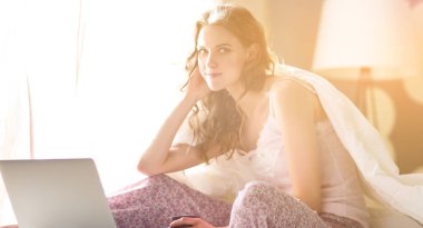 Dizüstü bilgisayar ile yatakta oturan genç güzel kadın.