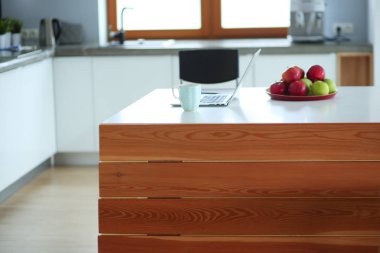 Genç kadın bir dizüstü bilgisayar kullanarak ve cep telefonuyla konuşurken mutfak masasında oturur.