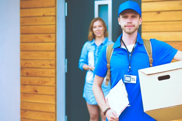 Χαμογελαστός διανομέας με μπλε στολή που παραδίδει δέματα στον παραλήπτη - έννοια της υπηρεσίας ταχυμεταφορών. Χαμογελαστός ντελιβεράς με μπλε στολή — Φωτογραφία Αρχείου
