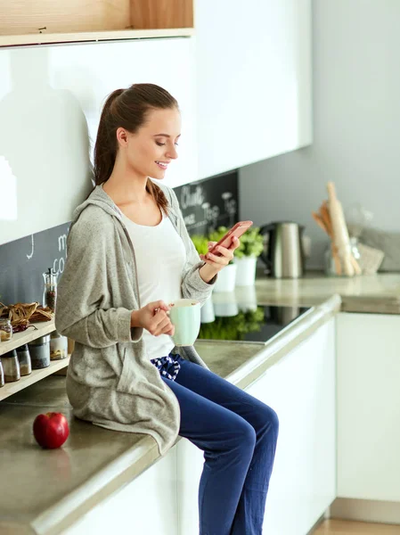 Mujer usando el teléfono móvil sentado en la cocina moderna . — Foto de Stock