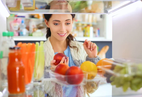 女性站在开着的冰箱旁边的画像，冰箱里装满了健康的食物、蔬菜和水果。女性的肖像 — 图库照片