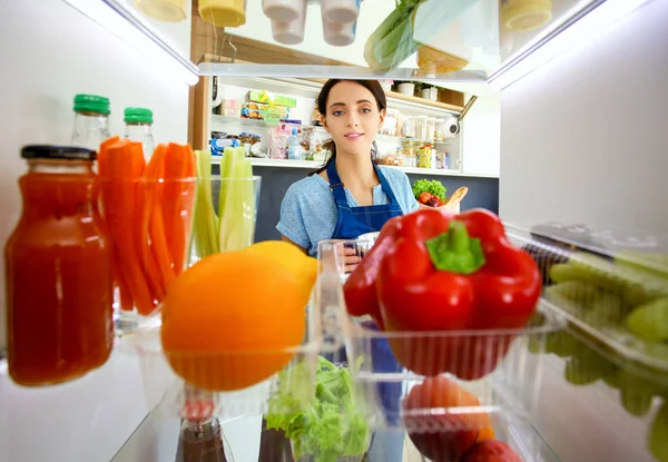 รูปภาพของผู้หญิงยืนอยู่ใกล้ตู้เย็นเปิดเต็มไปด้วยอาหารเพื่อสุขภาพผักและผลไม้ รูปภาพของผู้หญิง — ภาพถ่ายสต็อก