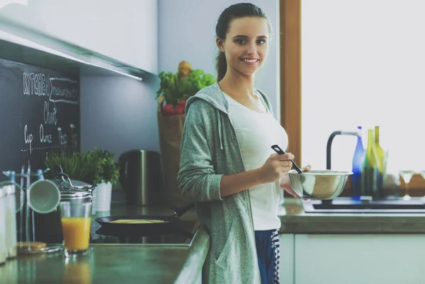 Eine junge Frau bereitet in der Küche Pfannkuchen zu, während sie am Tisch steht. Frau in der Küche. Kochen in der Küche. — Stockfoto