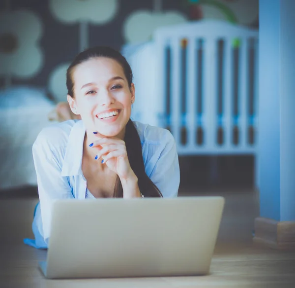 Giovane donna seduta sul pavimento vicino a lettino per bambini con computer portatile. Giovane mamma — Foto Stock