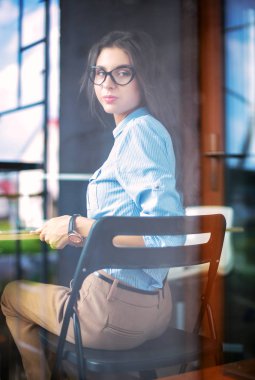 Ofis masasında dizüstü bilgisayarıyla oturan genç bir kadın. Pencereden bakıyor. Genç kadın.