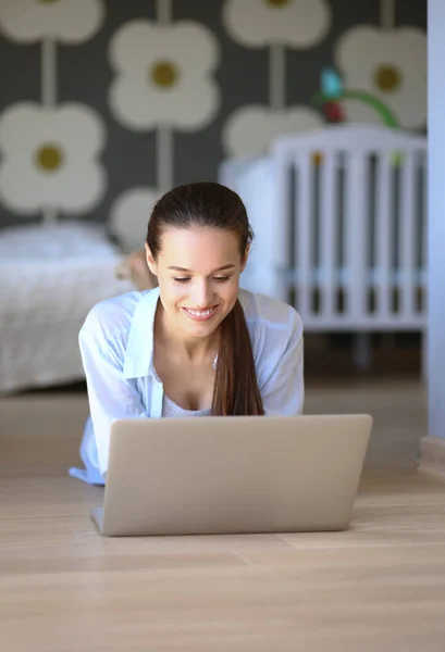 年轻的女人坐在笔记本电脑的儿童床边的地板上。年轻的妈妈 — 图库照片