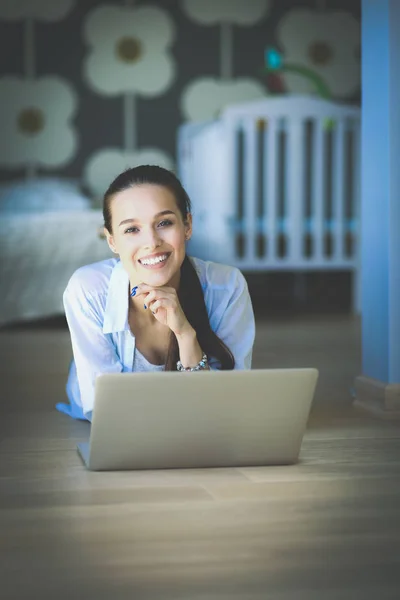 Ung kvinna som sitter på golvet nära barnens babysäng med laptop. Ung mamma — Stockfoto