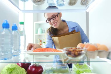 Sağlıklı yiyecek, sebze ve meyvelerle dolu açık buzdolabının yanında duran kadın portresi.