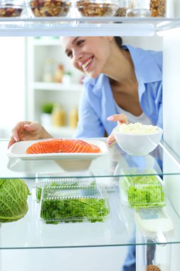 Sağlıklı yiyecek, sebze ve meyvelerle dolu açık buzdolabının yanında duran kadın portresi.