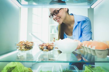 Sağlıklı yiyecek, sebze ve meyvelerle dolu açık buzdolabının yanında duran kadın portresi.
