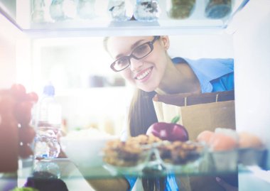 Sağlıklı yiyecek, sebze ve meyvelerle dolu açık buzdolabının yanında duran kadın portresi.