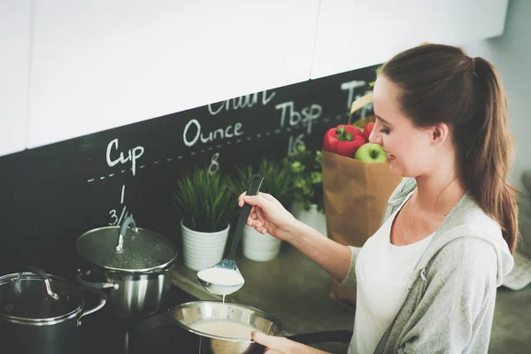 Jovem prepara panquecas na cozinha enquanto está perto da mesa. Uma mulher na cozinha. Cozinhar na cozinha. — Fotografia de Stock