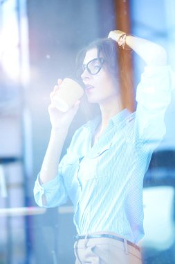 Ofis masasında dizüstü bilgisayarıyla oturan genç bir kadın. Pencereden bakıyor. Genç kadın.