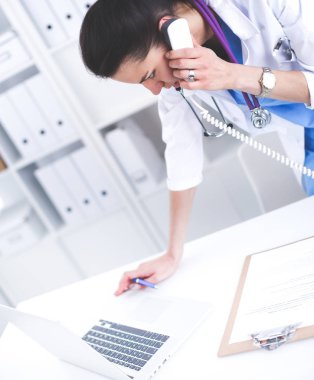 Bilgisayarda telefon kullanan beyaz önlüklü genç kadın doktor.