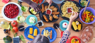 Ahşap masada otururken birlikte yemek yiyen bir grup insanın üst düzey görüntüsü. Masada yemek var. İnsanlar fast food yiyor..