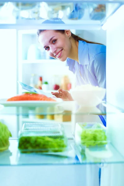 Retrato de fêmea em pé perto da geladeira aberta cheia de alimentos saudáveis, legumes e frutas — Fotografia de Stock