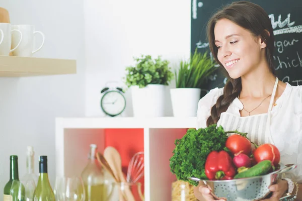 Mujer joven sonriente sosteniendo verduras de pie en la cocina. Jovencita sonriente —  Fotos de Stock