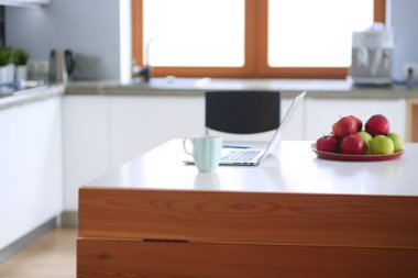 Genç kadın bir dizüstü bilgisayar kullanarak ve cep telefonuyla konuşurken mutfak masasında oturur.