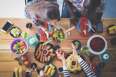 Ahşap masada otururken birlikte yemek yiyen bir grup insanın üst düzey görüntüsü. Masada yemek var. İnsanlar fast food yiyor..