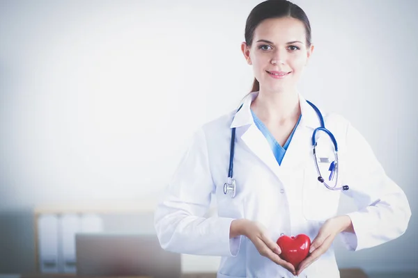 Vackra unga leende kvinnliga läkare sitter vid skrivbordet och hålla hjärtat. — Stockfoto