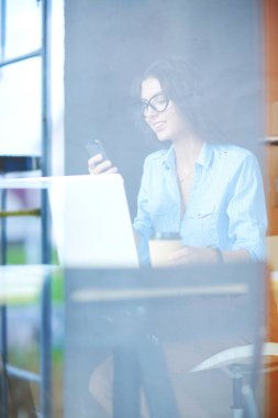 Ofis masasında dizüstü bilgisayarıyla oturan genç bir kadın. Pencereden bakıyor. Genç kadın.