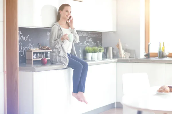 Mujer usando el teléfono móvil sentado en la cocina moderna . — Foto de Stock