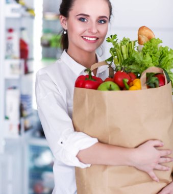 Sebzeli alışveriş torbası tutan genç bir kadın. Mutfakta duruyor. Mutfakta kameraya bakan bir kadın var.
