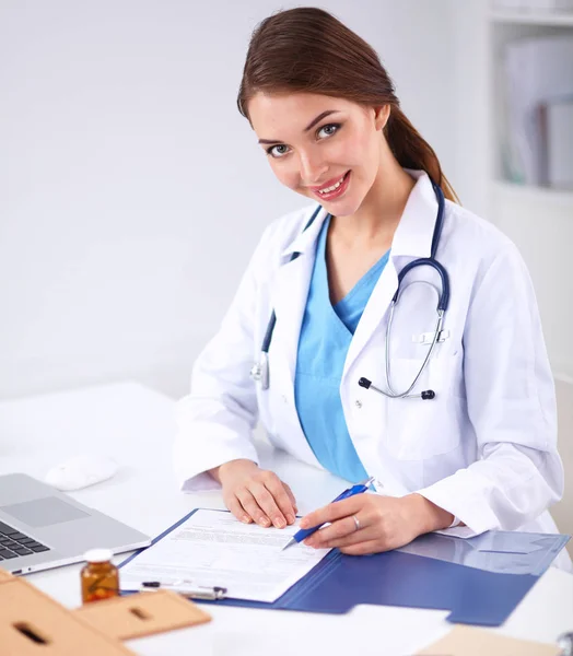 Hermosa joven sonriente doctora sentada en el escritorio y escribiendo. — Foto de Stock