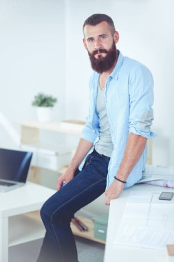 Modern bir ofiste tek başına dururken bilgisayar masasına yaslanan gülümseyen genç bir tasarımcının portresi.