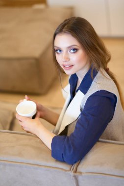 Genç bir kadın, beyaz odada rahat bir koltukta, elinde kahveyle oturuyor.