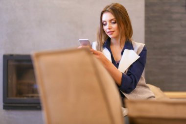 Genç bir kadın, beyaz odada rahat bir koltukta, elinde kahveyle oturuyor.