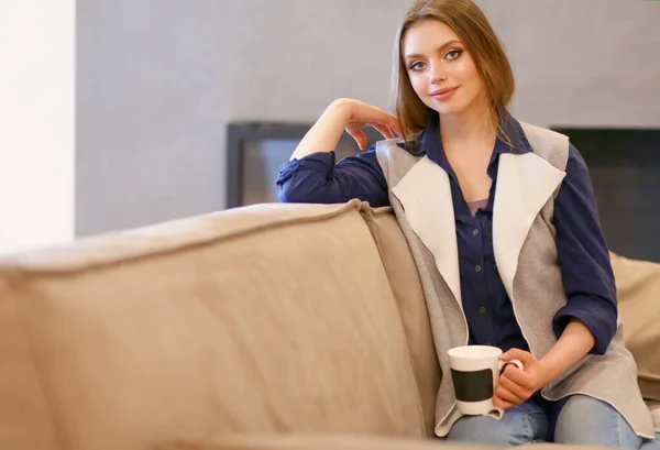 Ung kvinna sitter på bekväm soffa med kopp på kaffe i händerna i vitt rum — Stockfoto