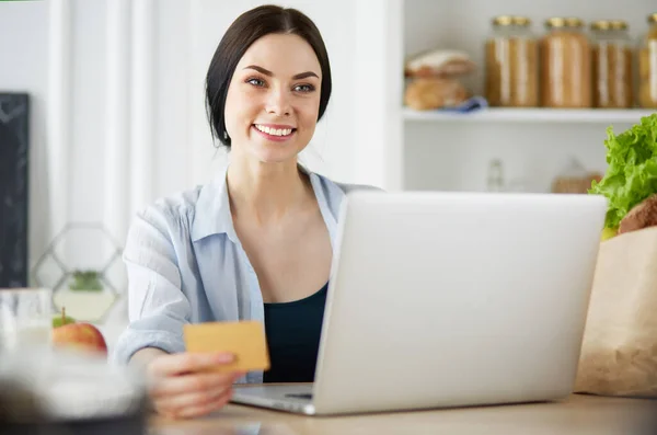 Mulher sorrindo compras on-line usando computador e cartão de crédito na cozinha — Fotografia de Stock