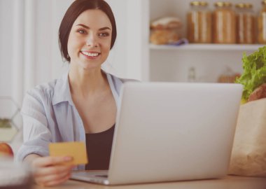Mutfakta bilgisayar ve kredi kartı kullanan gülümseyen bir kadın.