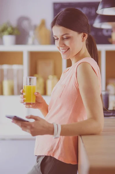 Lachende mooie vrouw kijken naar mobiele telefoon en glas sinaasappelsap te houden terwijl het ontbijten in een keuken. — Stockfoto