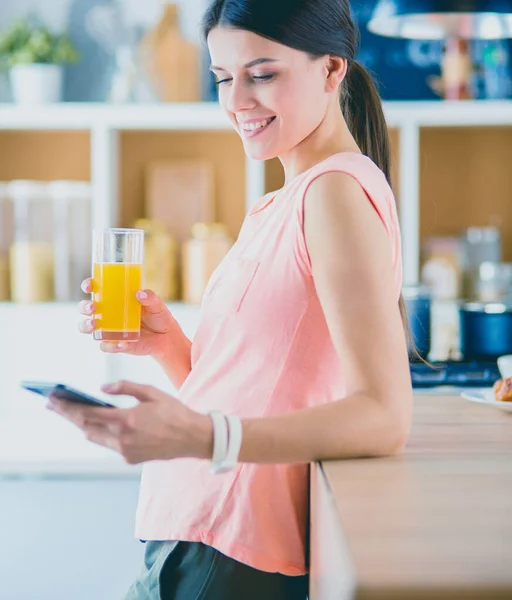 Mujer bonita sonriente mirando el teléfono móvil y sosteniendo un vaso de jugo de naranja mientras desayunaba en una cocina . — Foto de Stock