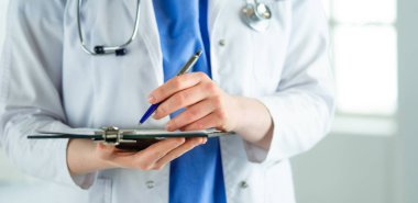Steteskoplu bir doktor elinde bir defter tutuyor. Hastanede ayakta dikilirken panoda tıbbi form dolduran bir kadın doktorun yakın çekimi.