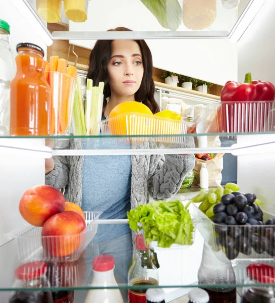 Portrait of cute serious female standing near open fridge full of healthy food, vegetables and fruits — Stock Photo, Image