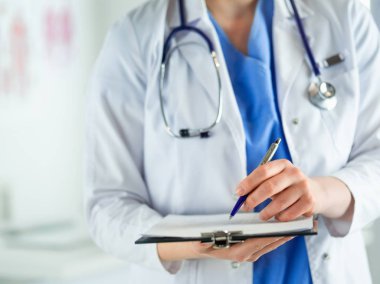 Steteskoplu bir doktor elinde bir defter tutuyor. Hastanede ayakta dikilirken panoda tıbbi form dolduran bir kadın doktorun yakın çekimi.