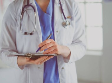 Steteskoplu bir doktor elinde bir defter tutuyor. Hastanede ayakta dikilirken panoda tıbbi form dolduran bir kadın doktorun yakın çekimi.