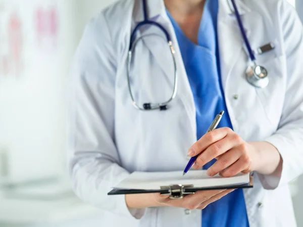 청진기를 들고, 손에 공책을 들고 있는 의사. 병원에서 똑바로 서 있는 한 여의사가 클립보드에서 의학적 서류를 작성하는 모습 — 스톡 사진