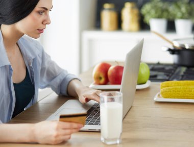 Mutfakta bilgisayar ve kredi kartı kullanan gülümseyen bir kadın.