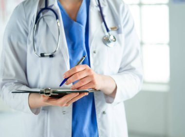 Steteskoplu bir doktor elinde bir defter tutuyor. Hastanede ayakta dikilirken panoda tıbbi form dolduran bir kadın doktorun yakın çekimi.