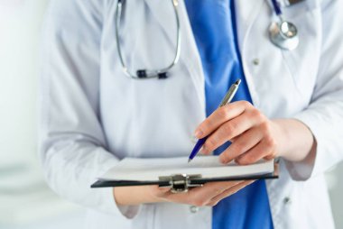 Steteskoplu bir doktor elinde bir defter tutuyor. Hastanede ayakta dikilirken panoda tıbbi form dolduran bir kadın doktorun yakın çekimi.